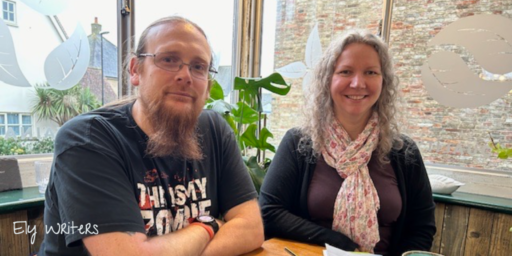 Left to right: Greg and Caity. Greg and Caity are sitting in the corner of a cafe with a plant in the window behind them and notebooks on the table in front. In the bottom left corner, it says 'Ely Writers'.