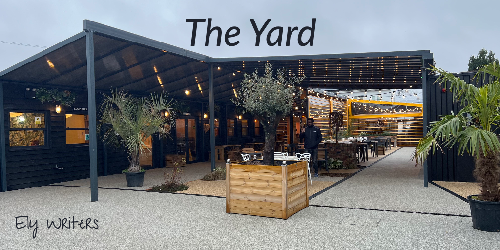 Photo of The Yard by Silver Oak Coffee. There is a yard surrounded on two sides with enclosed areas. In the yard itself, there is seating and planters with little trees.